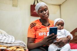 A Kenyan woman looks at a smartphone while holding a child in her lap